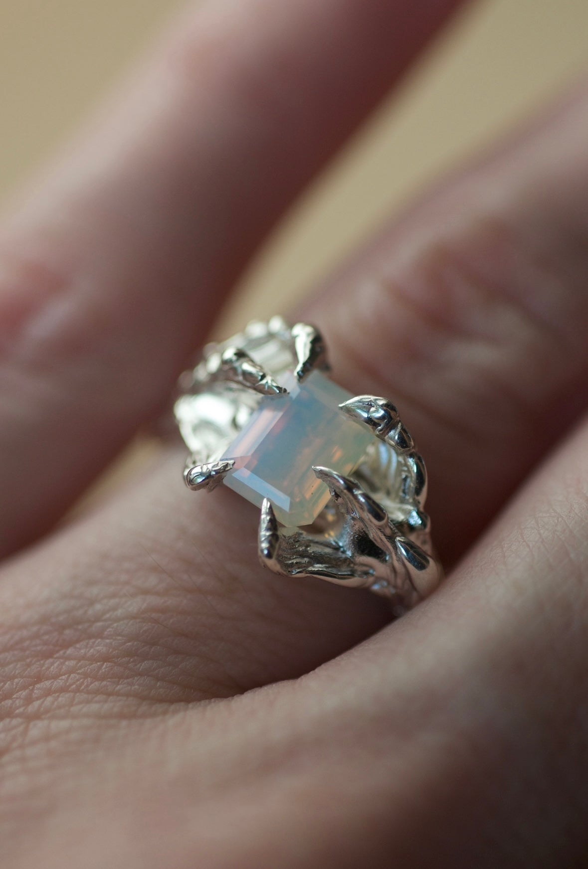 Lost wax cast sterling silver ring with organic and molten features. The centre stone is a milky opal emerald cut lab grown crystal. The ring is sitting on a finger.